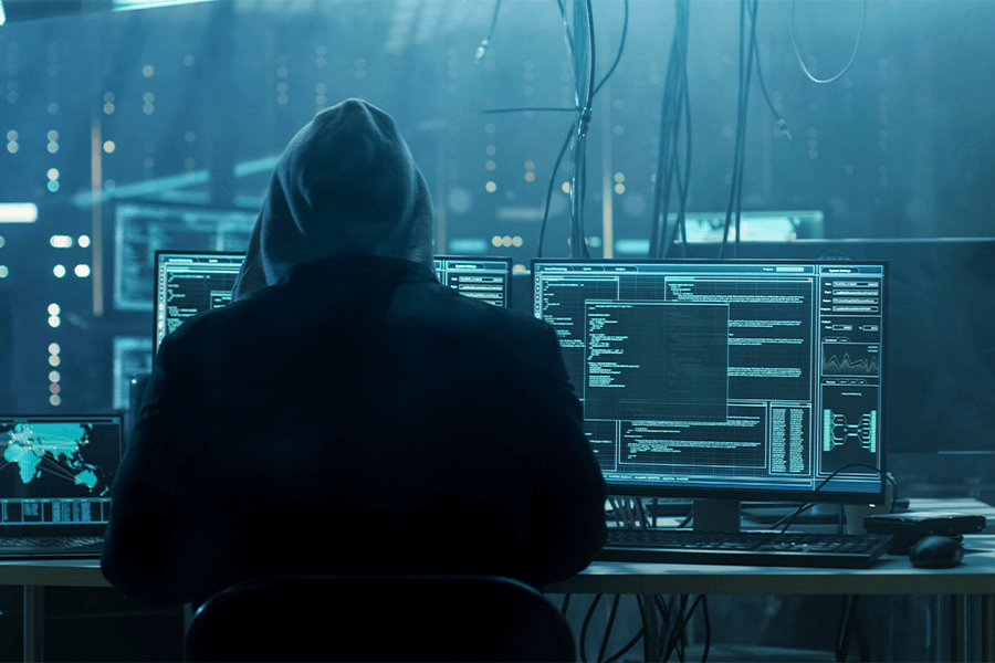 A hooded figure seen from behind at a desk of monitors showing code and dashboards in a darkened room